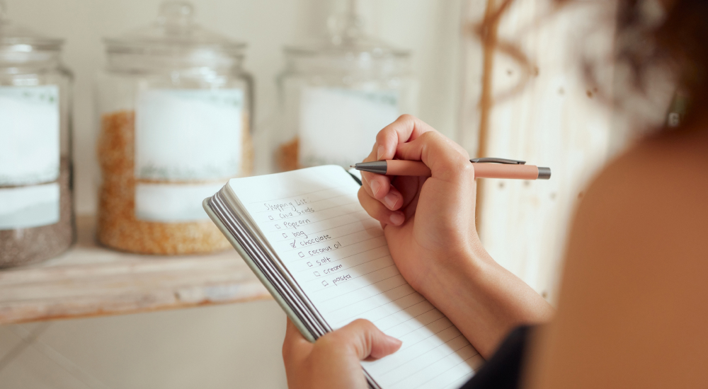 Image shows focus on a woman’s hands as she creates a shopping list in a notebook.